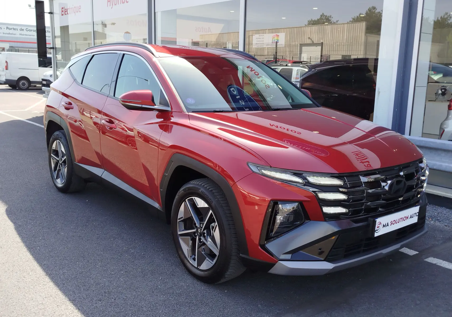 Hyundai Tucson 2024 hybride en Ultimate Red Métal, vu en 3/4 avant droit avec calandre noire et jantes bi-ton.