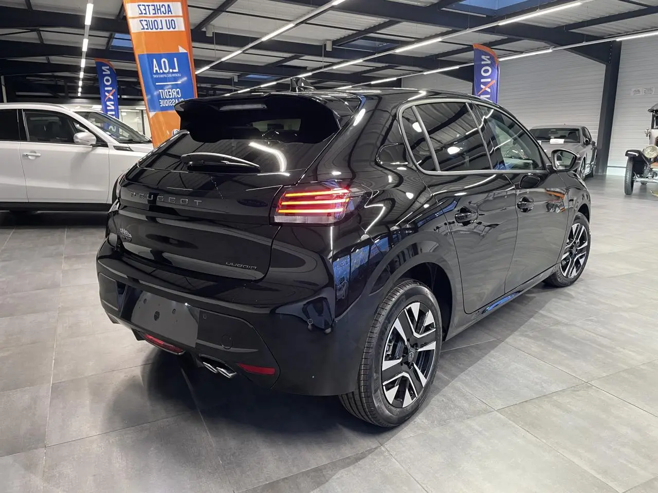 Vue 3/4 arrière droite d'une Peugeot 208 noire métallisée avec feux arrière allumés en showroom intérieur.