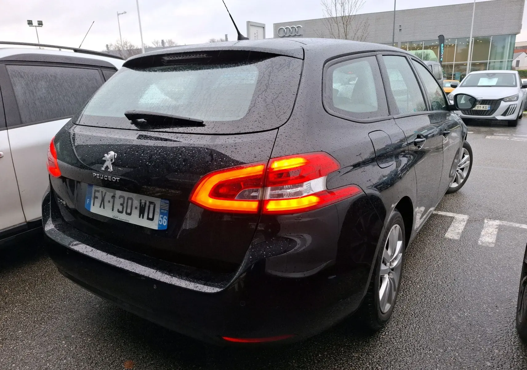 Peugeot 308 SW noire vue en 3/4 arrière droit sous la pluie avec feux arrière allumés sur parking.
