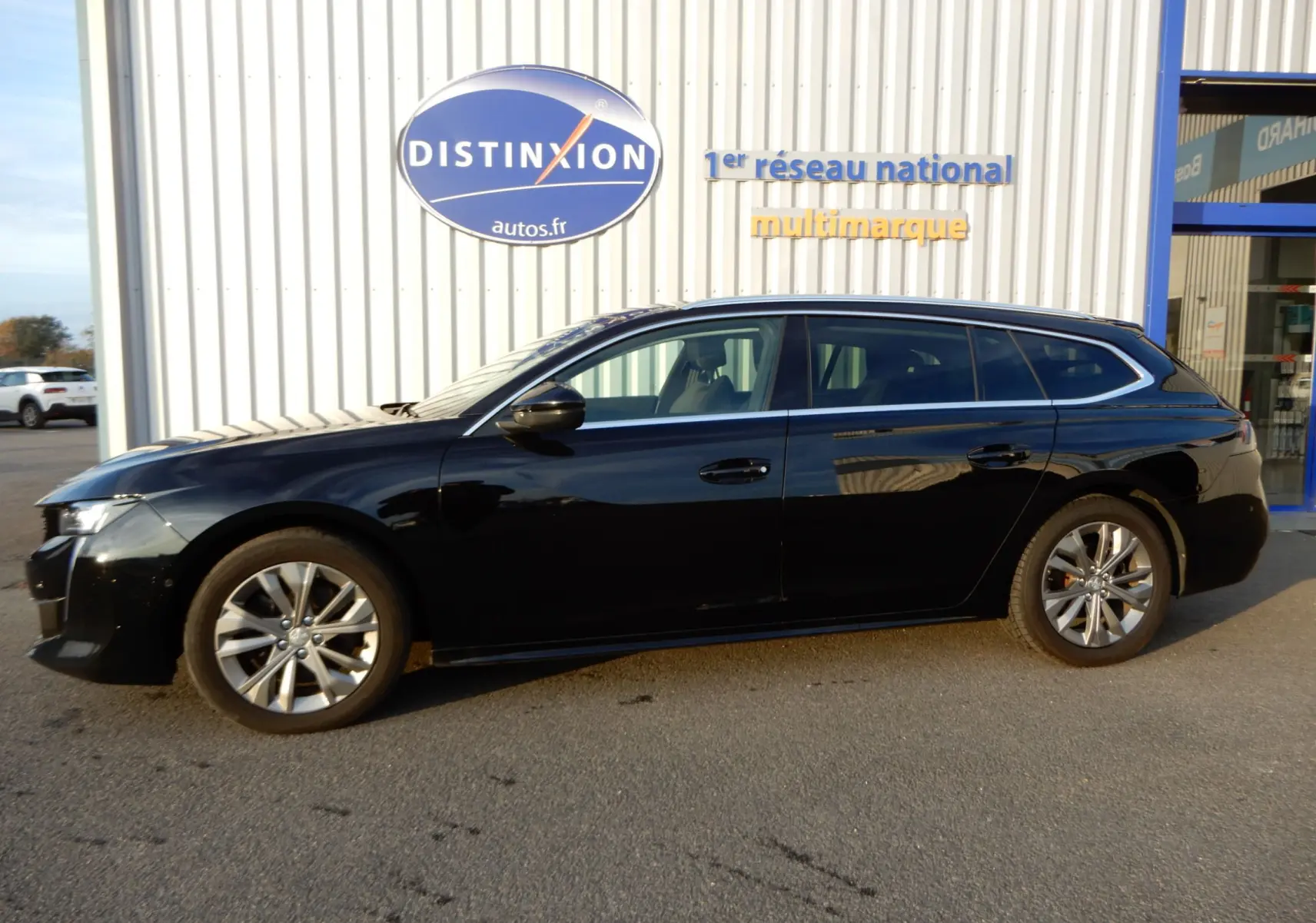 Peugeot 508 SW noir Perla Nera vue de profil côté gauche, stationnée devant un bâtiment avec enseigne Distinxion.