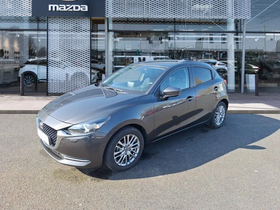 Vue 3/4 avant droit d'une Mazda2 gris métallisé garée devant un showroom Mazda avec jantes alliage.