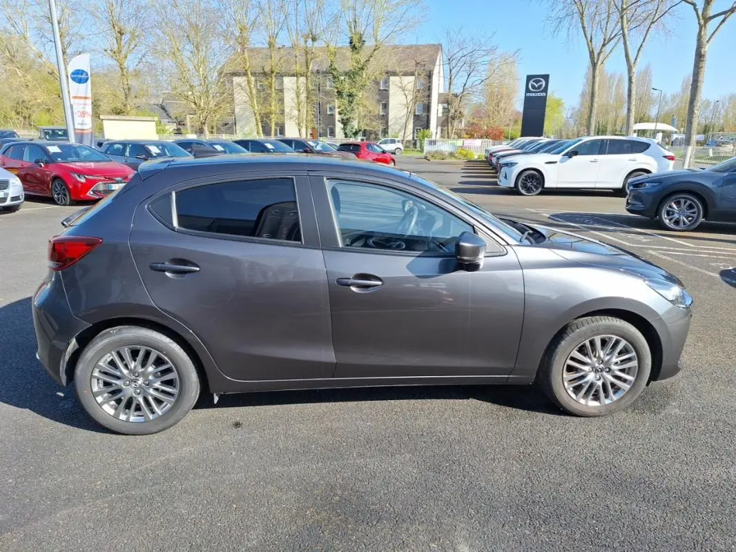 Profil côté gauche d'une Mazda2 gris métallisé 2023 avec jantes alliage et vitres surteintées sur parking extérieur.