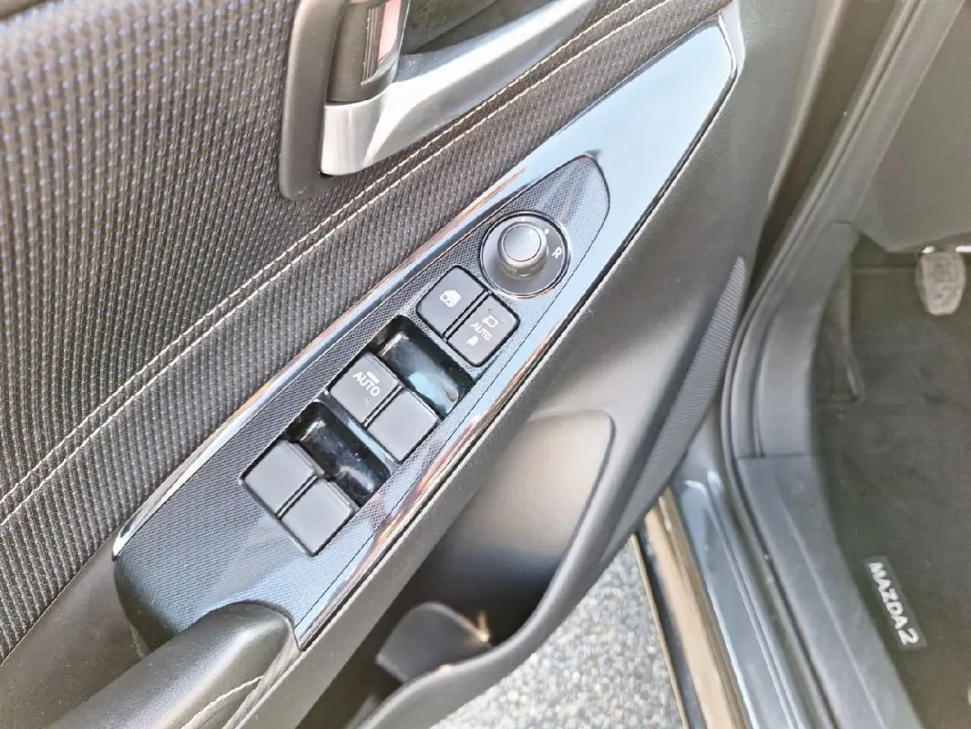 Vue rapprochée du panneau de commandes des vitres côté conducteur d'une Mazda2 gris 2023 avec boutons auto et réglage rétroviseurs.