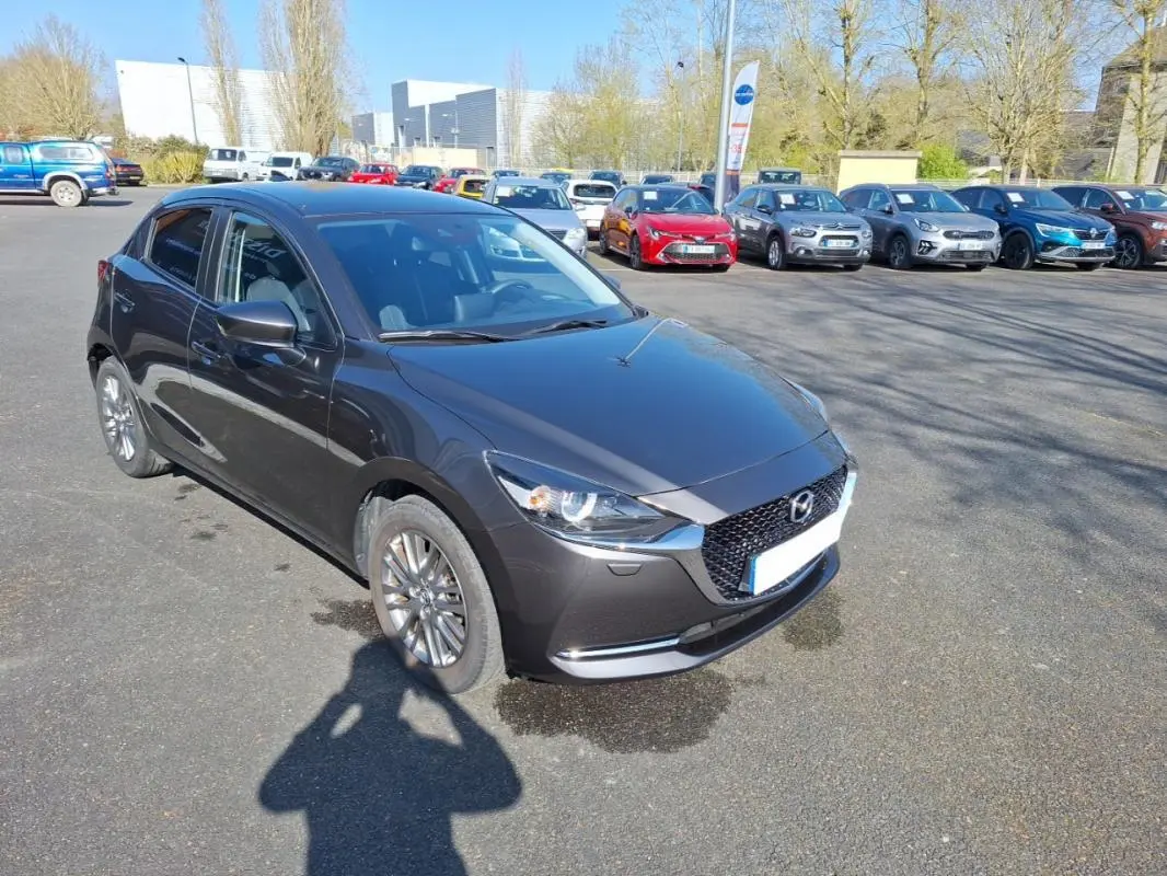 Vue 3/4 avant gauche d'une Mazda2 gris métallisé avec jantes aluminium sur un parking extérieur.