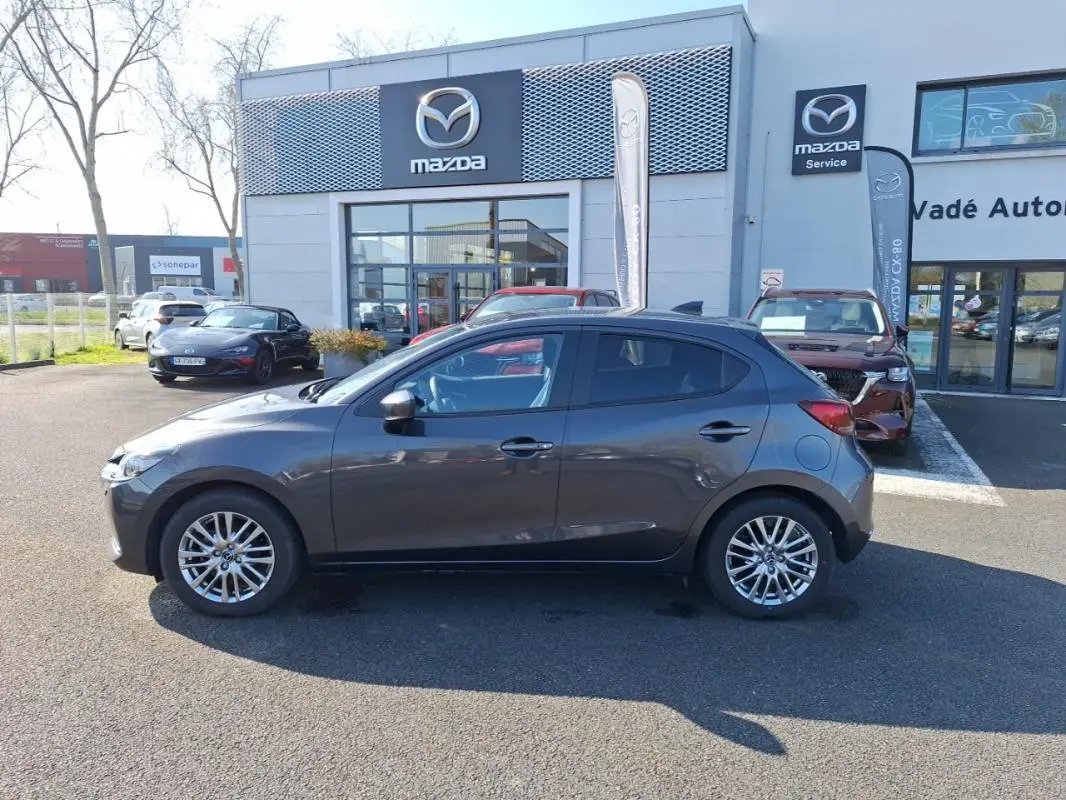 Vue de profil côté gauche d'une Mazda2 gris métallisé stationnée devant un concessionnaire Mazda.