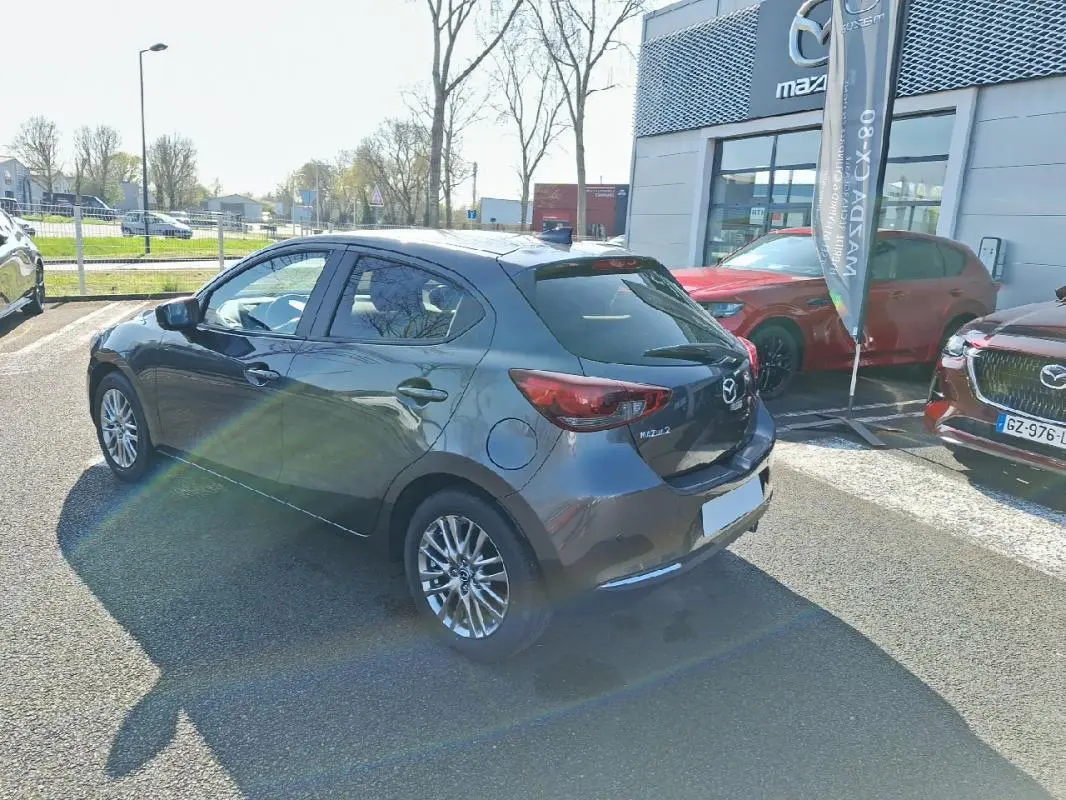 Vue 3/4 arrière droite d'une Mazda2 gris métallisé 2023 avec jantes aluminium, stationnée devant un concessionnaire Mazda.