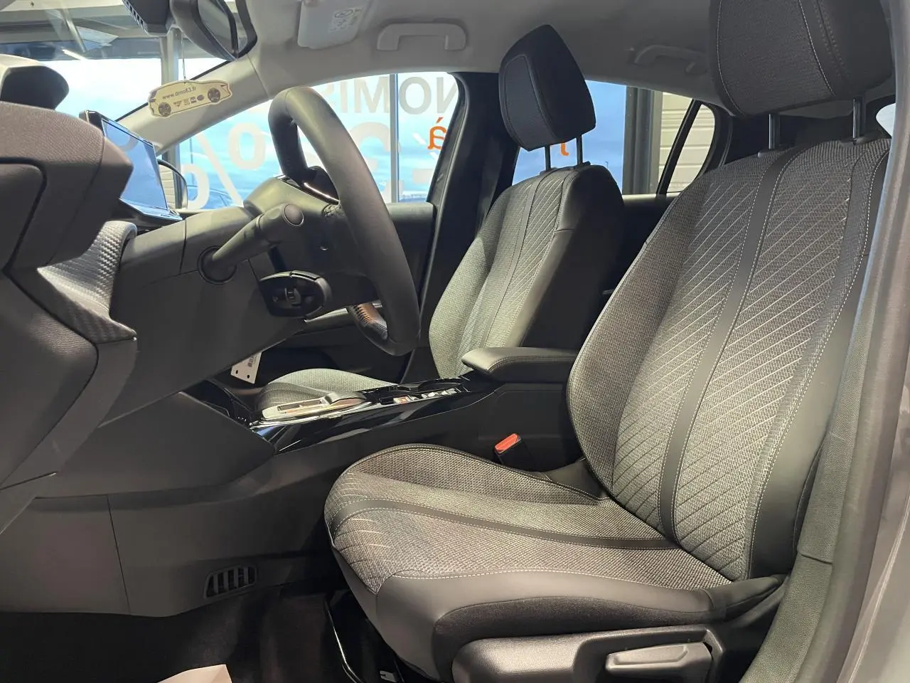 Vue intérieure côté gauche montrant les sièges avant gris clair et le volant de la Peugeot 208 hybride 2025.
