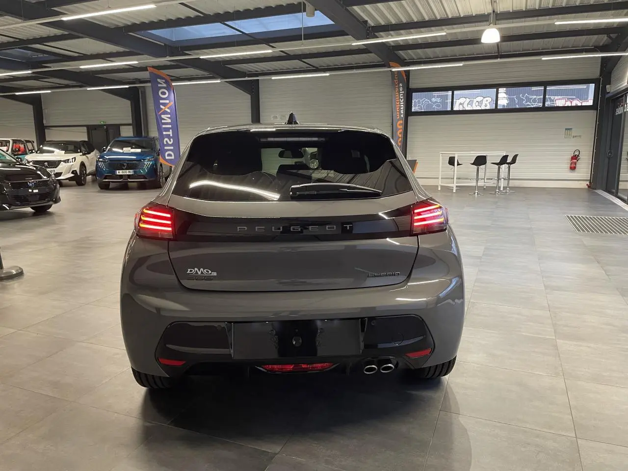 Vue arrière d'une Peugeot 208 hybride gris selenium avec feux arrière allumés en intérieur showroom.