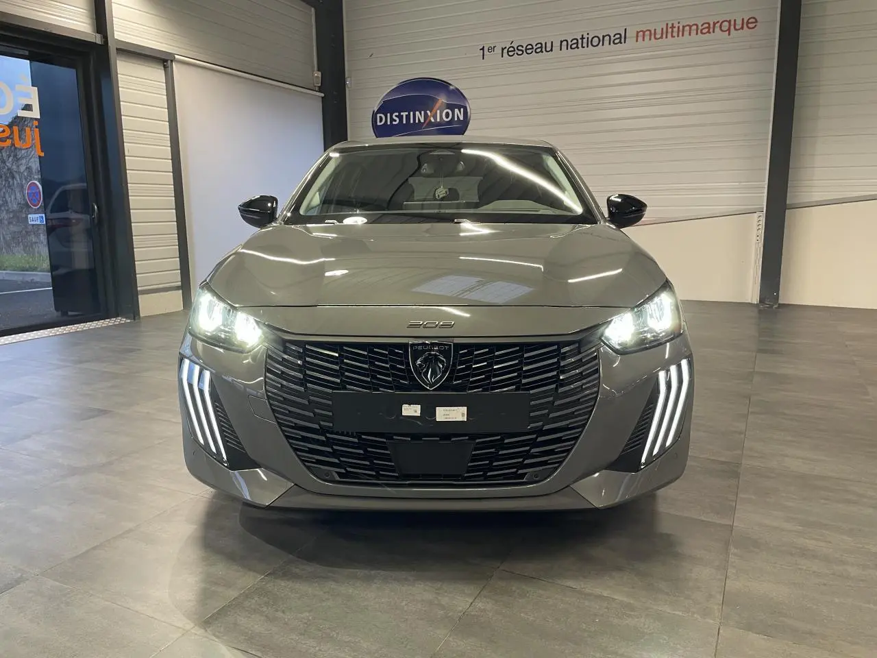 Vue avant d'une Peugeot 208 gris selenium avec phares allumés et signature lumineuse verticale LED visible en intérieur showroom.