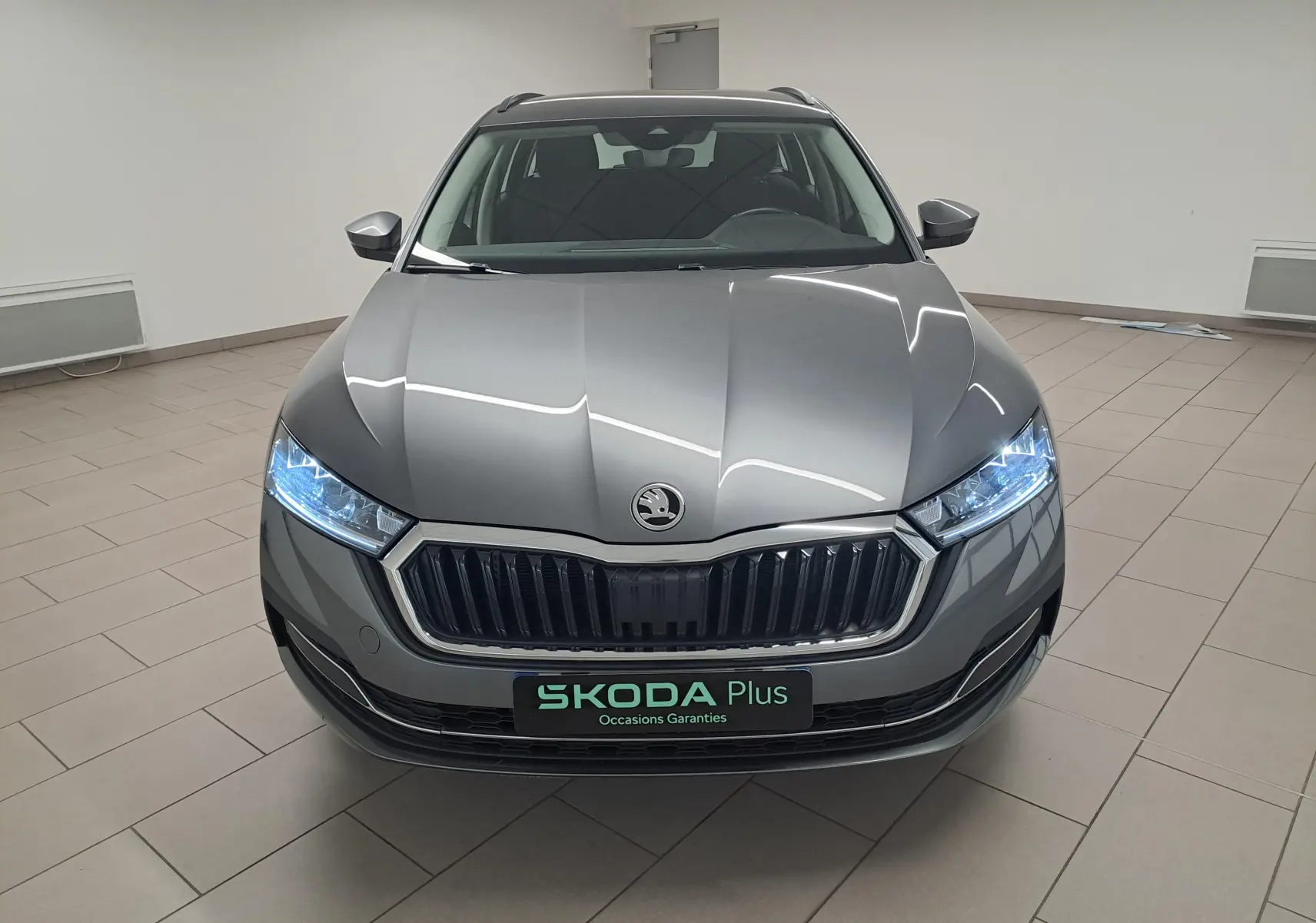 Vue avant d'une Skoda Octavia Combi 2024 gris graphite avec phares allumés dans un showroom.