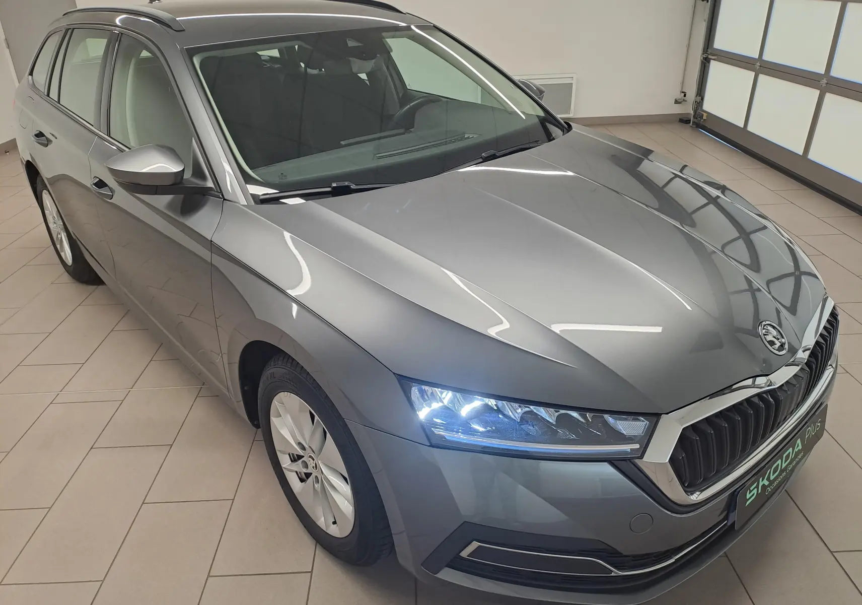 Vue 3/4 avant droit d'une Skoda Octavia Combi 2024 gris graphite avec phares allumés dans un garage.