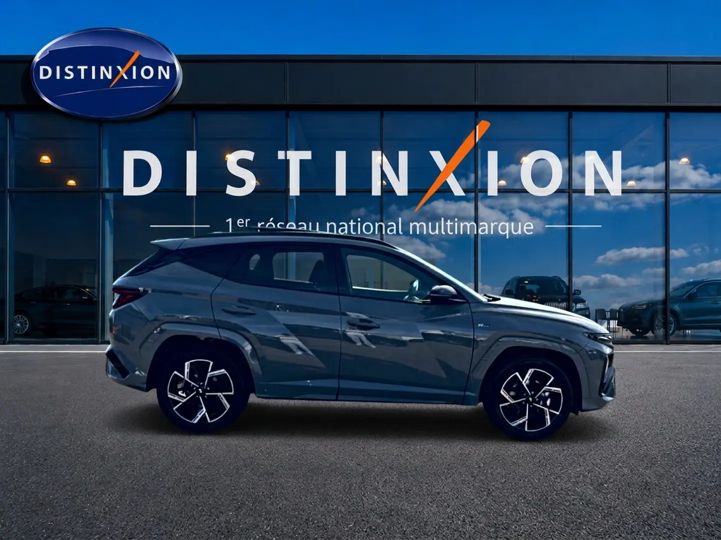 Hyundai Tucson 2026 gris shadow en profil gauche devant vitrine Distinxion, avec jantes alu et lignes marquées.