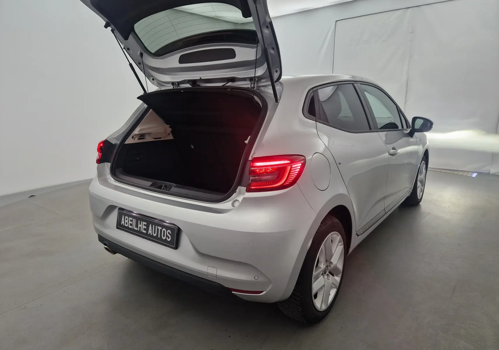 Vue 3/4 arrière droite d'une Renault Clio V gris platine 2021 avec coffre ouvert et feux arrière allumés.