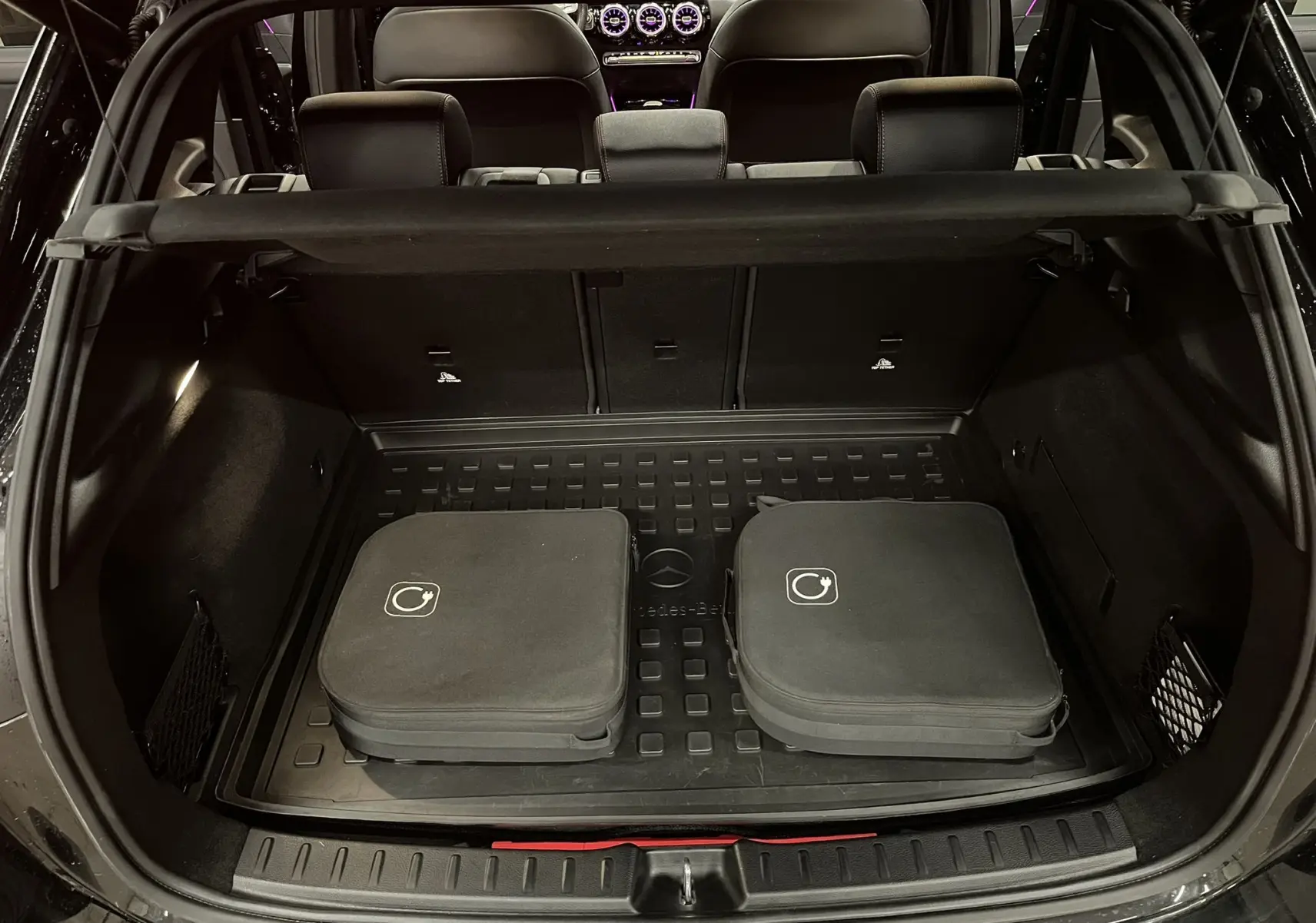 Coffre ouvert d’un Mercedes GLA 250E AMG Line noir Cosmos, avec tapis de coffre caoutchouc et deux housses noires.