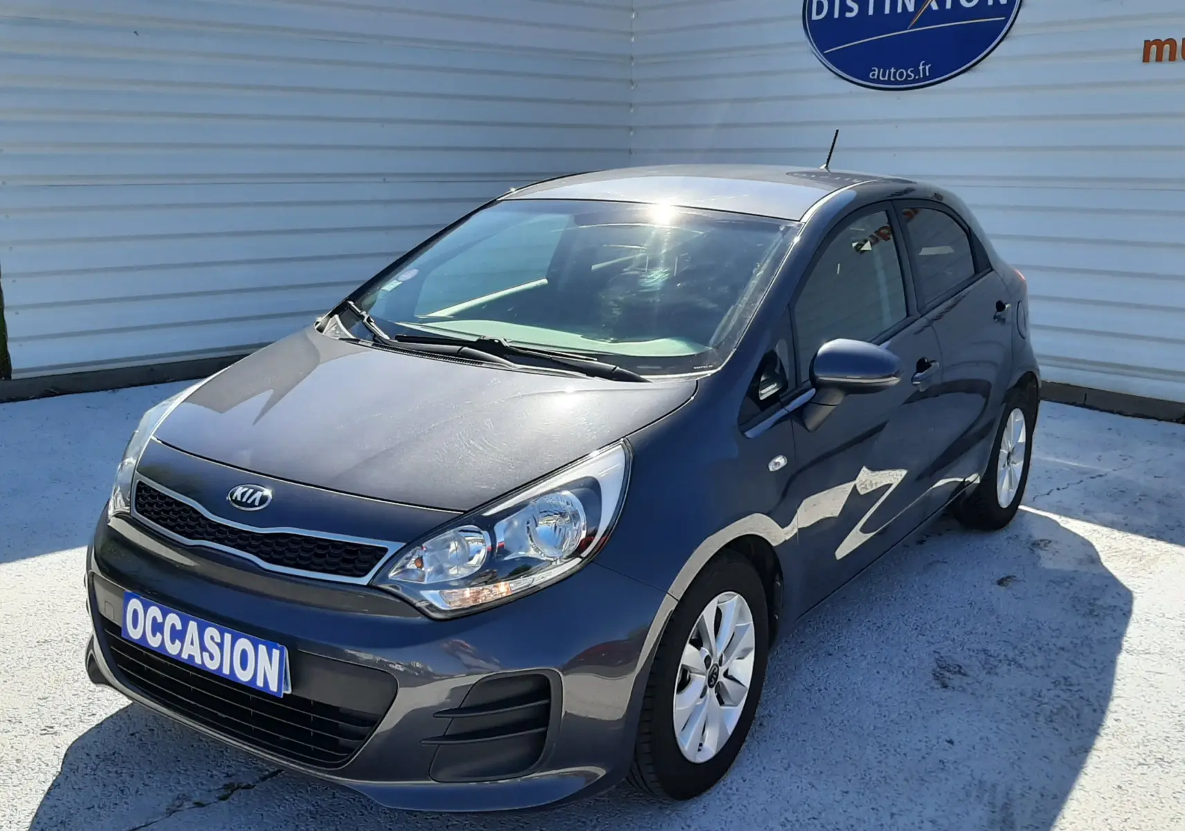 KIA Rio grise vue 3/4 avant droit, compacte 5 portes avec jantes alliage et plaque "OCCASION" visible.