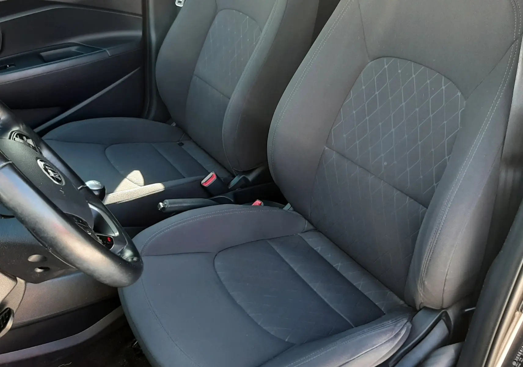 Vue intérieure côté conducteur de la Kia Rio 2016, sièges tissu gris avec motifs losanges et volant visible.