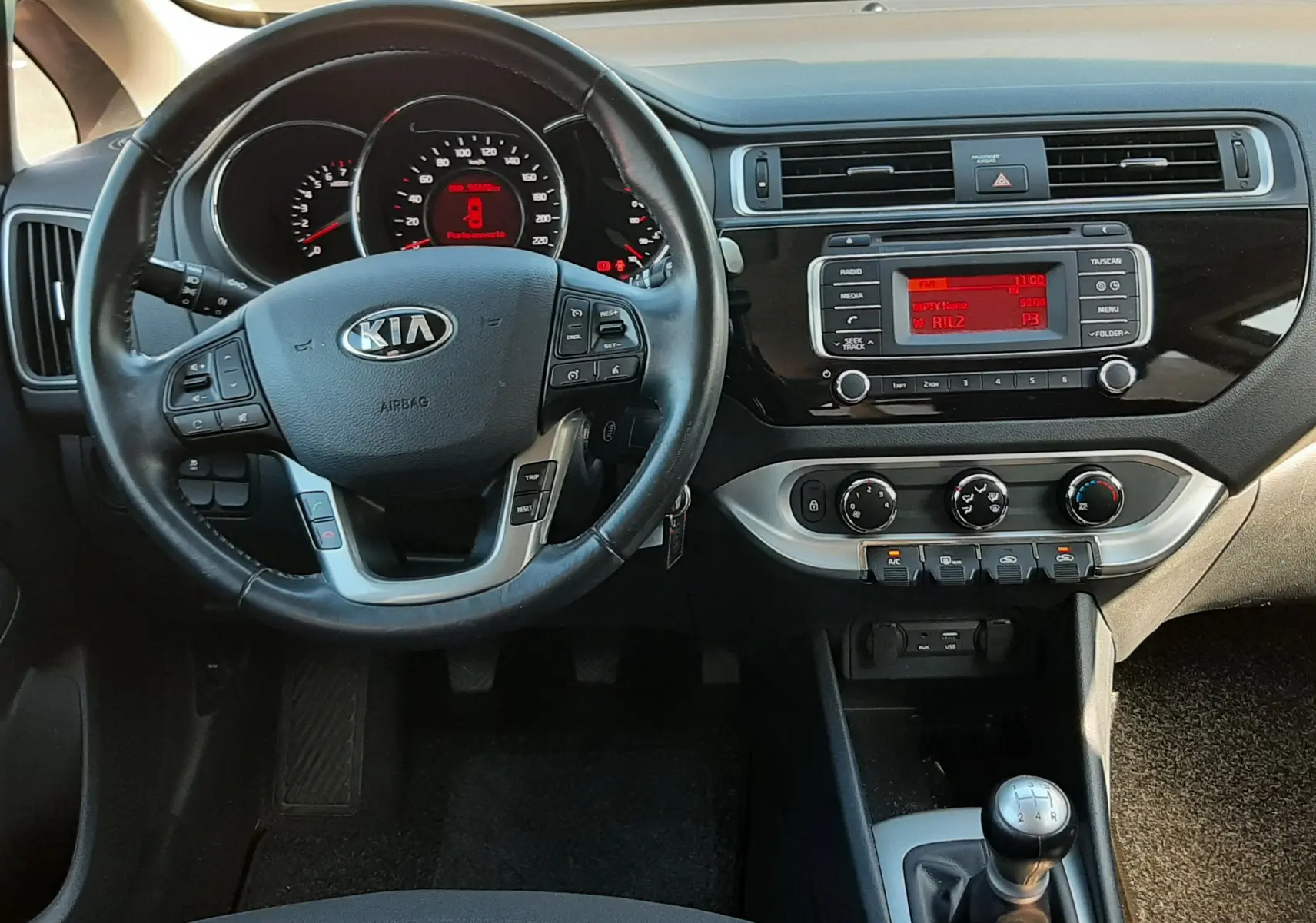 Vue intérieure frontale du tableau de bord et volant noir de la KIA RIO grise 2016 avec boîte manuelle et écran radio central.