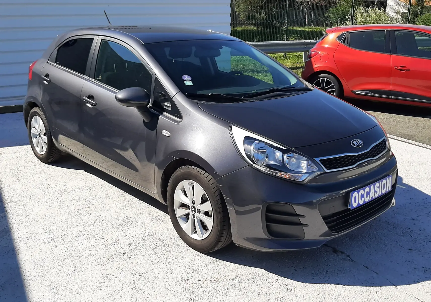 KIA Rio grise vue 3/4 avant droit, compacte 5 portes avec jantes alliage et plaque "OCCASION" visible.
