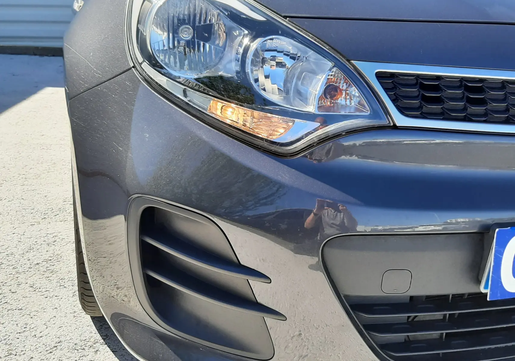 Gros plan sur l'avant droit d'une KIA Rio grise 2016, mettant en valeur le phare et la calandre noire.