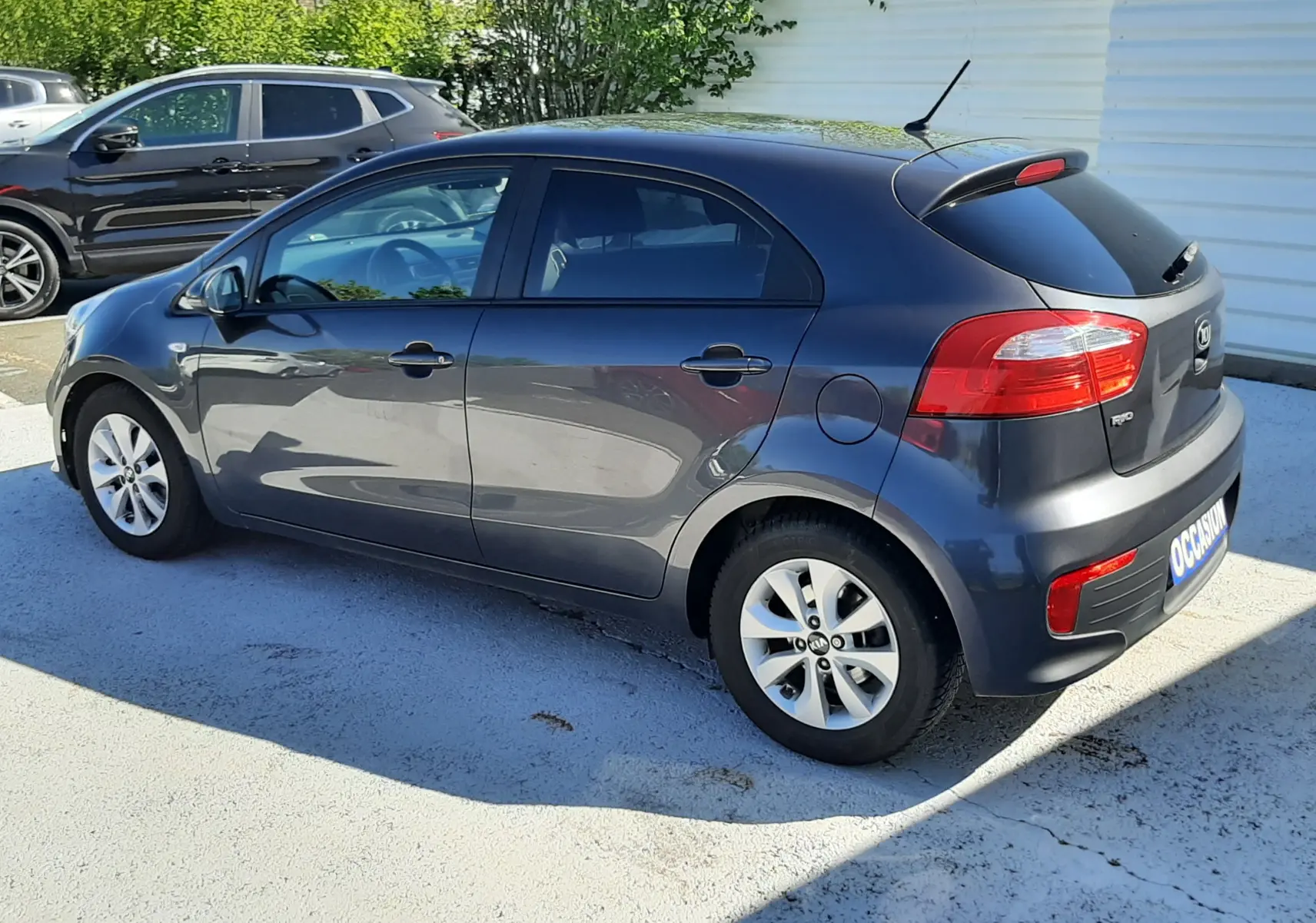 KIA Rio grise vue 3/4 arrière côté gauche, compacte 5 portes avec jantes alliage et feux arrière rouges.