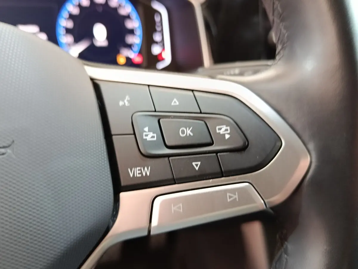 Gros plan sur les commandes multifonctions du volant cuir alu du Volkswagen Taigo gris foncé, tableau de bord numérique en arrière-plan.