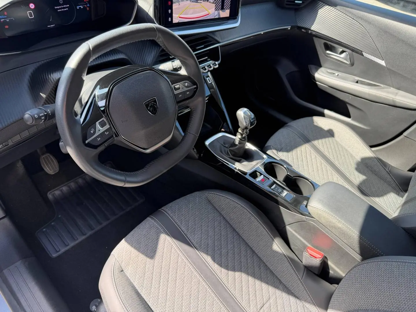 Vue intérieure côté conducteur de la Peugeot 208 1.2 100ch avec volant compact, sellerie grise et boîte manuelle.