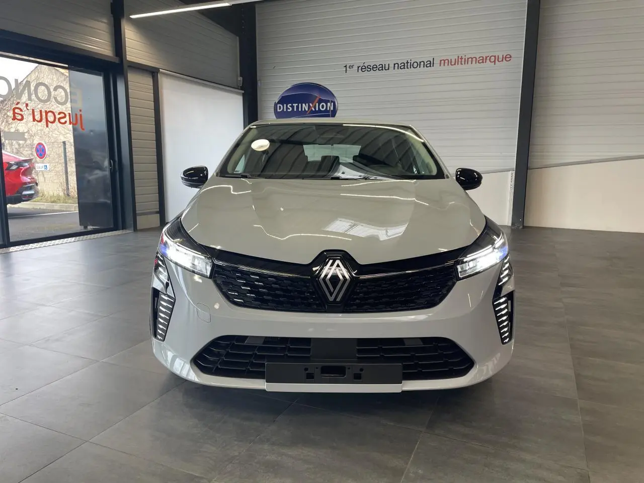 Renault Clio 1.5 Blue dCi 2025 en gris Rafale vue de face en intérieur, avec calandre noire et feux LED allumés.