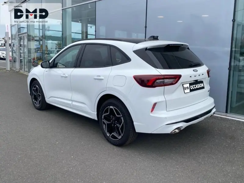 Vue 3/4 arrière droite d'un Ford Kuga blanc Glacier avec jantes alu noires et toit panoramique, stationné devant un bâtiment vitré.