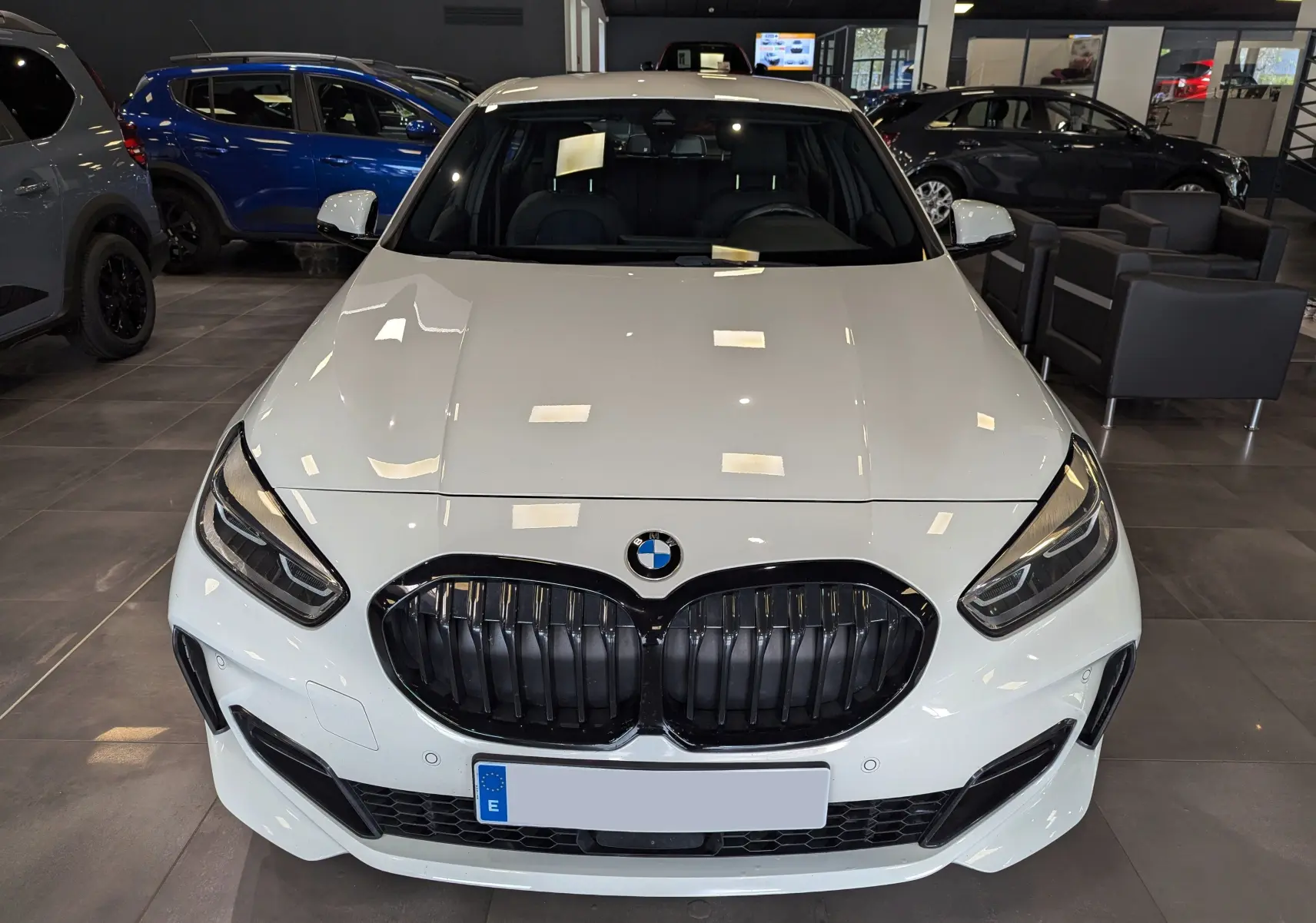 Vue frontale d'une BMW Série 1 F40 18d blanche Alpine avec calandre noire et phares LED dans un showroom.