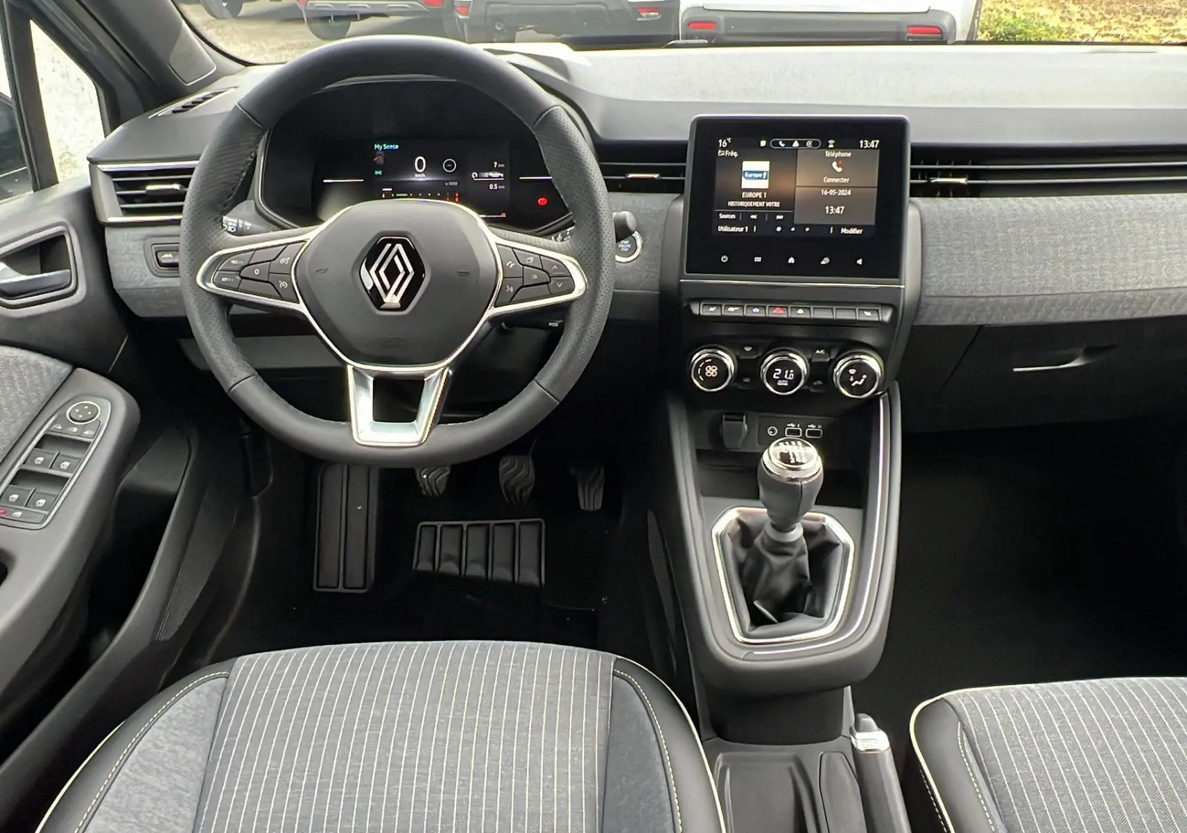 Intérieur de la Renault Clio V TCE 90 TECHNO 2025, vue du poste de conduite avec volant, tableau de bord digital et levier de vitesse manuel.