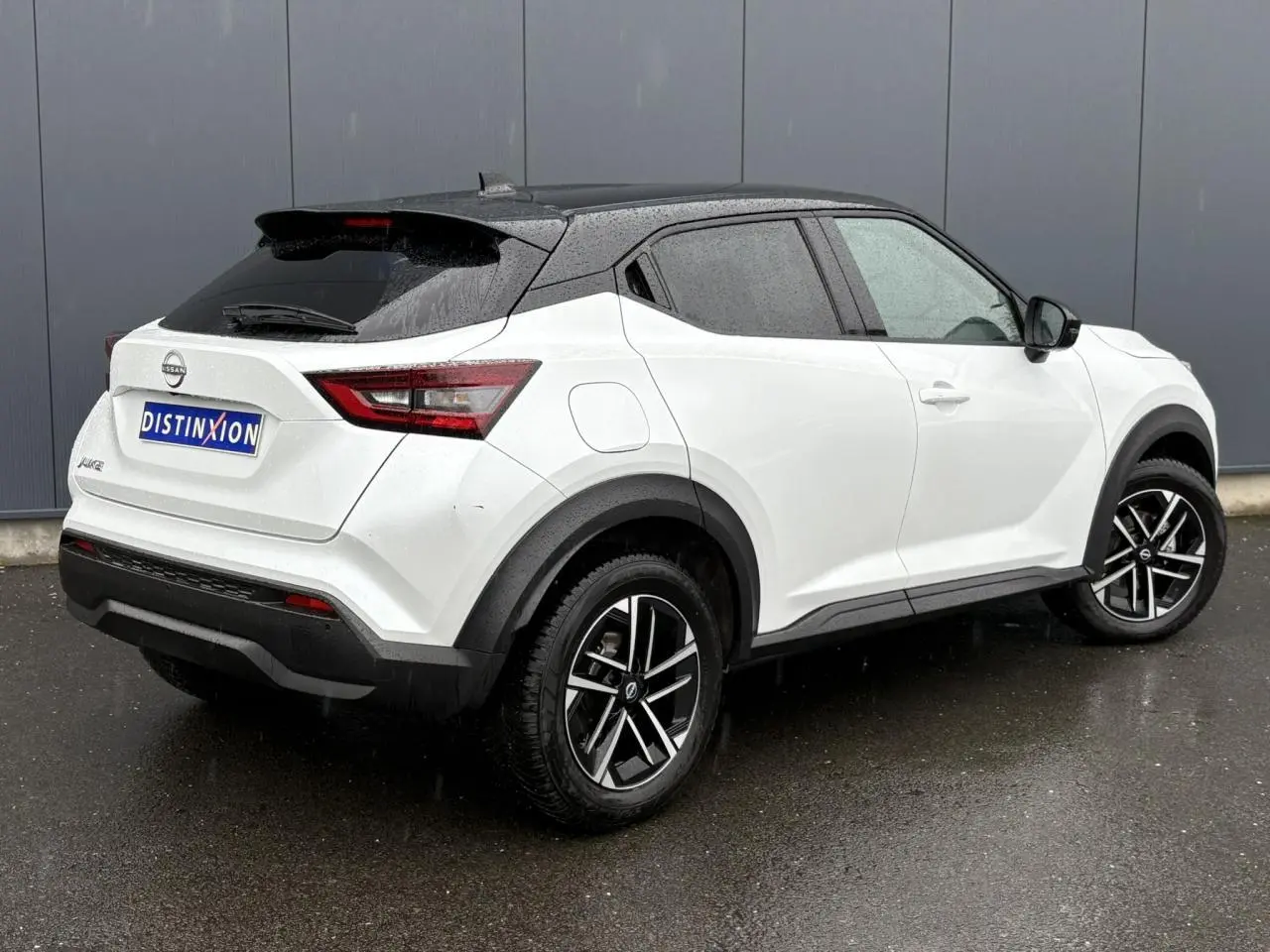 Nissan Juke blanc nacré vue 3/4 arrière droit sous la pluie avec toit noir et jantes alliage 17 pouces.