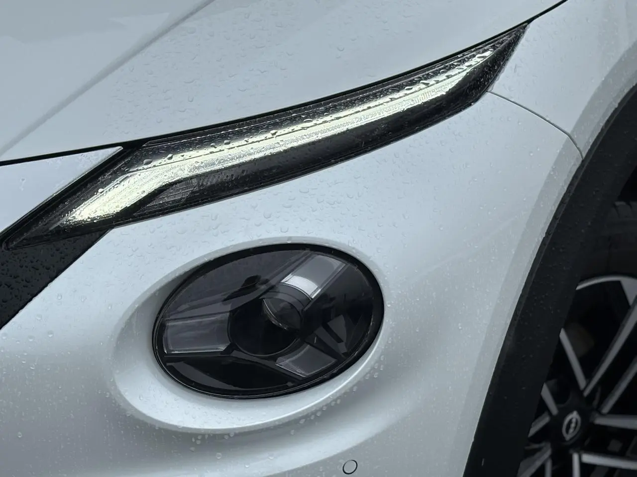Gros plan sur le phare avant droit et la jante alliage du Nissan Juke blanc nacré sous la pluie.