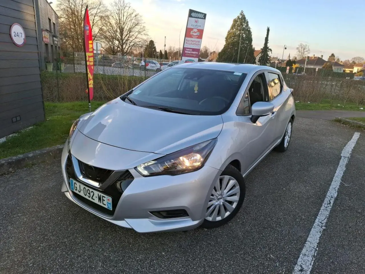 Nissan Micra gris clair vue 3/4 avant droit avec phares allumés, stationnée sur un parking extérieur.
