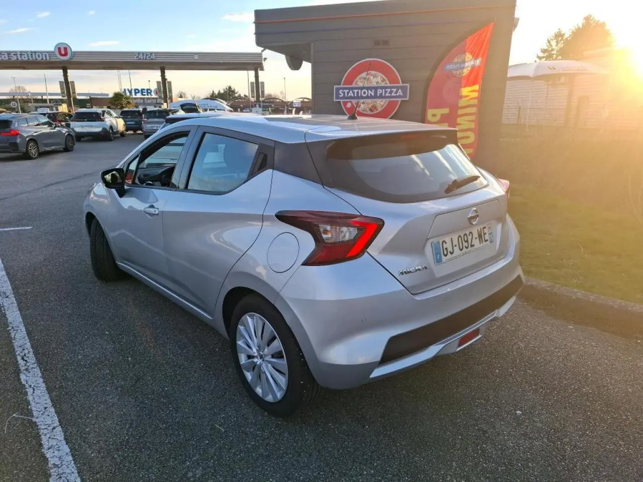 Vue 3/4 arrière droite d'une Nissan Micra gris clair garée près d'une station-service au coucher du soleil.