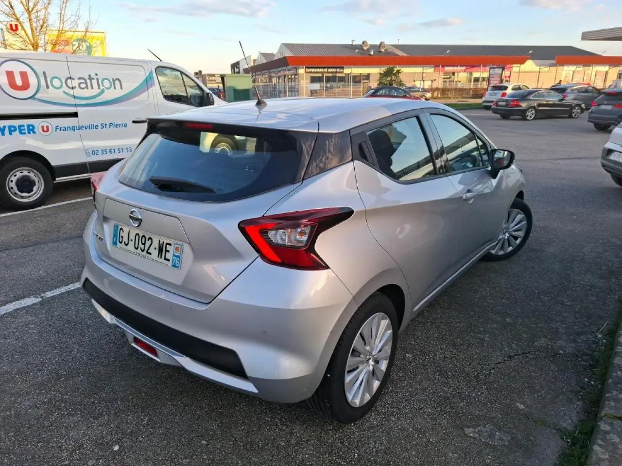 Vue 3/4 arrière droite d'une Nissan Micra gris clair avec jantes en acier et spoiler arrière, stationnée en extérieur.