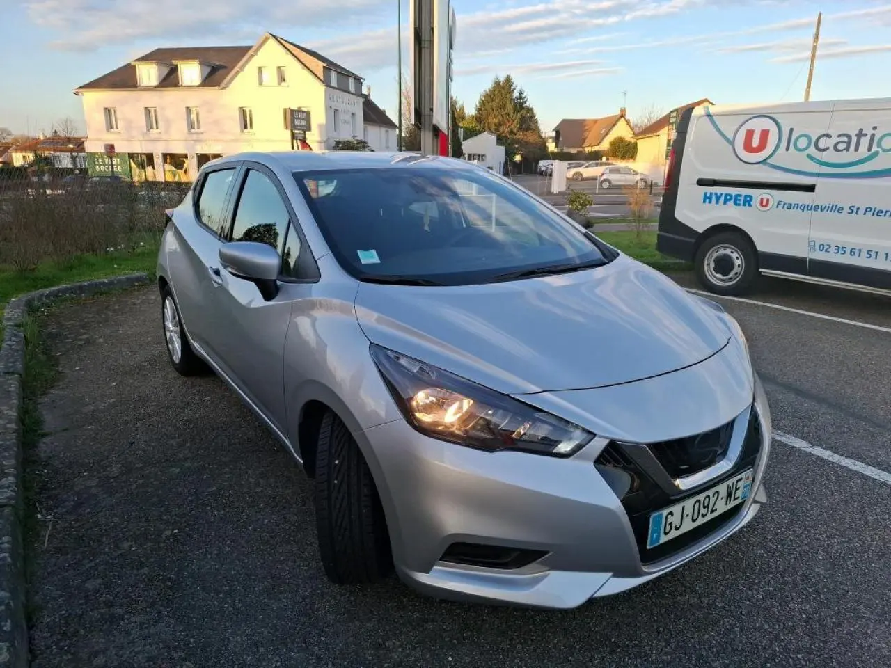 Nissan Micra gris clair vue 3/4 avant droit, phare avant allumé, stationnée sur un parking extérieur.