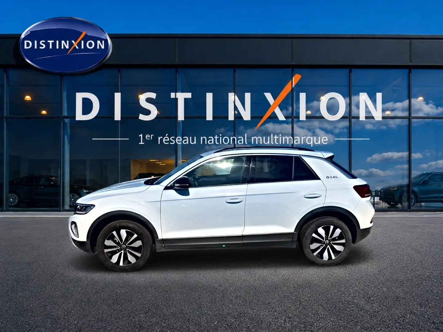 Volkswagen T-Roc blanc en profil droit, avec jantes alliage 17 pouces et barres de toit visibles, devant une vitrine Distinxion.