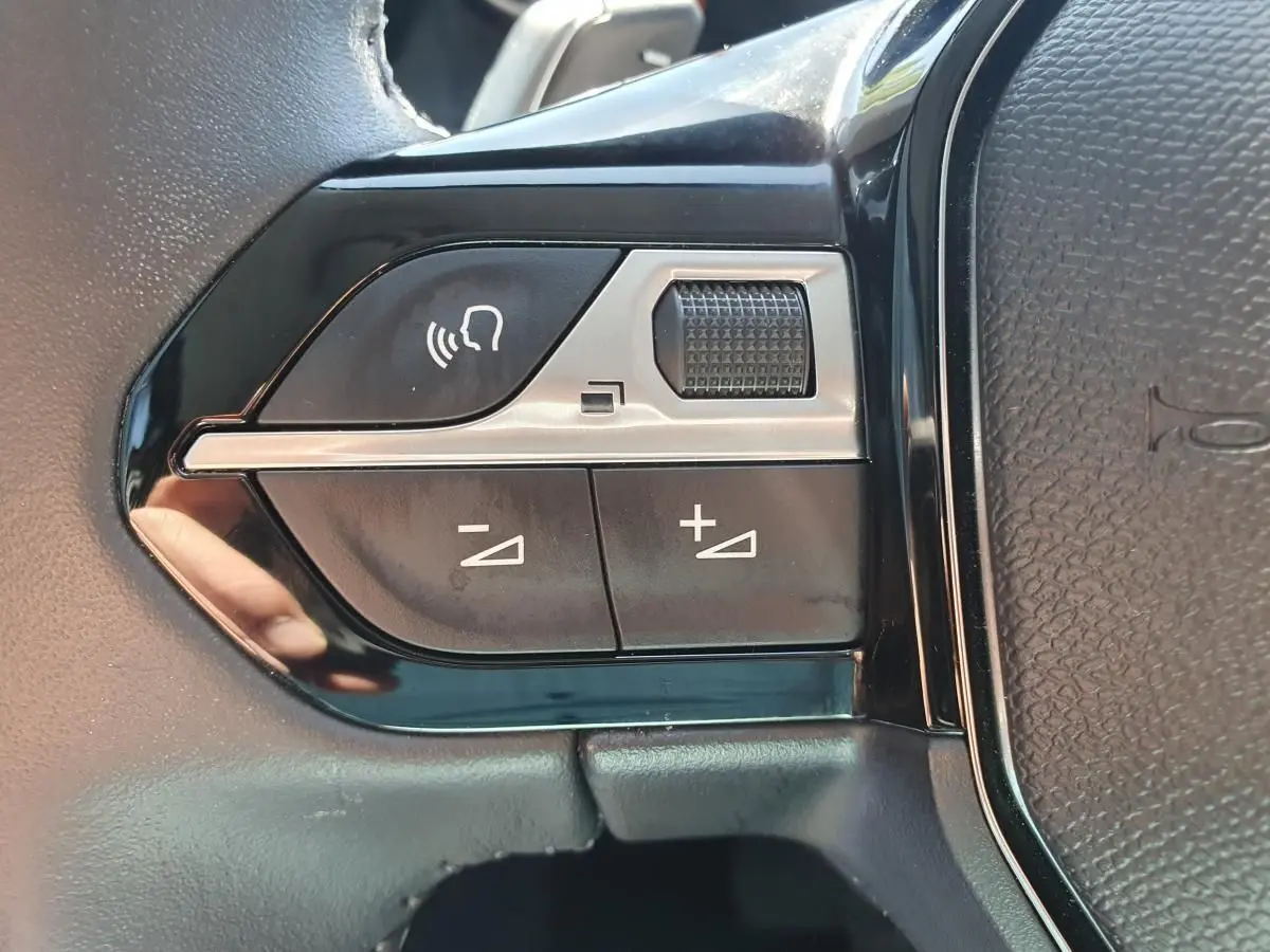 Gros plan sur les commandes de volume vocal et sonore au volant du Peugeot 208 gris Artense, version hybride 2025.