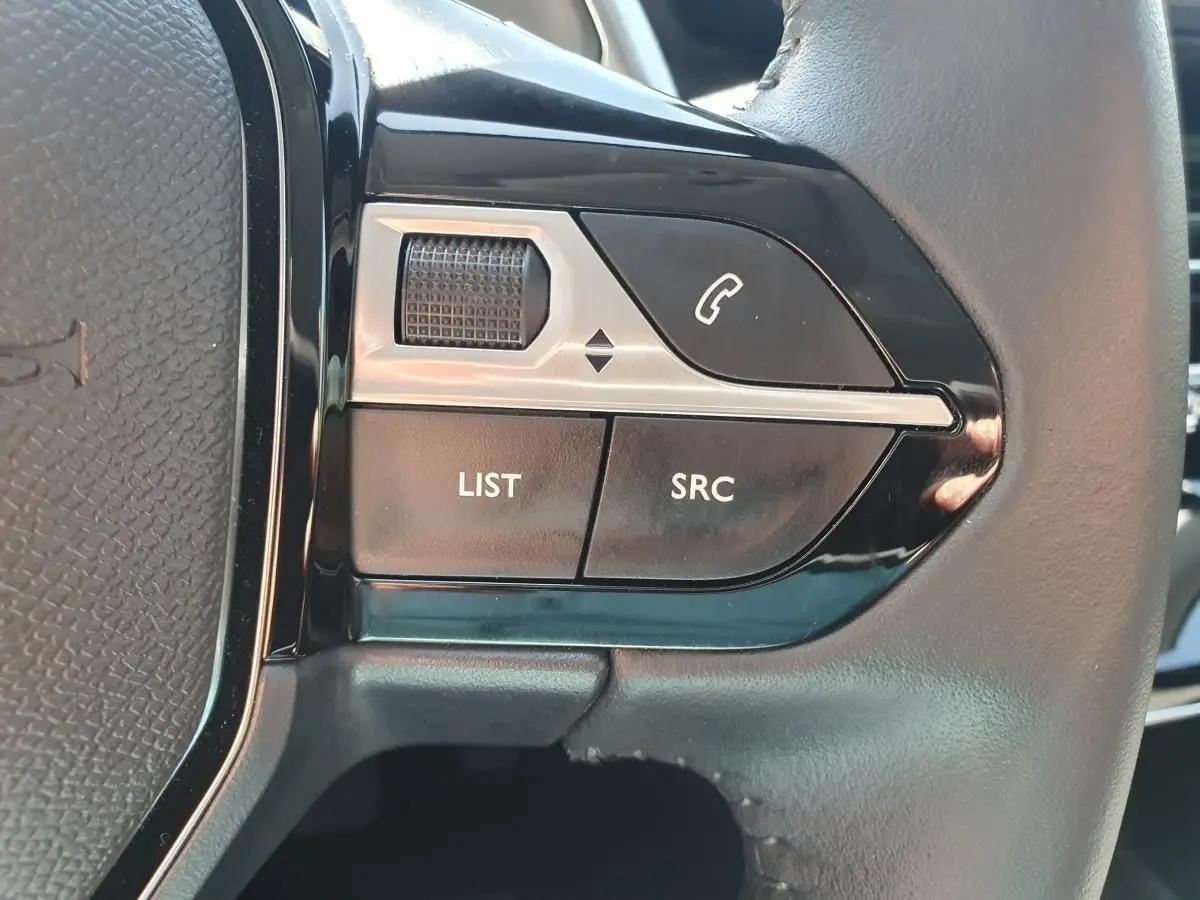 Gros plan sur les commandes gauche du volant noir de la Peugeot 208 1.2 Hybrid 110 gris Artense, boutons appel et audio.