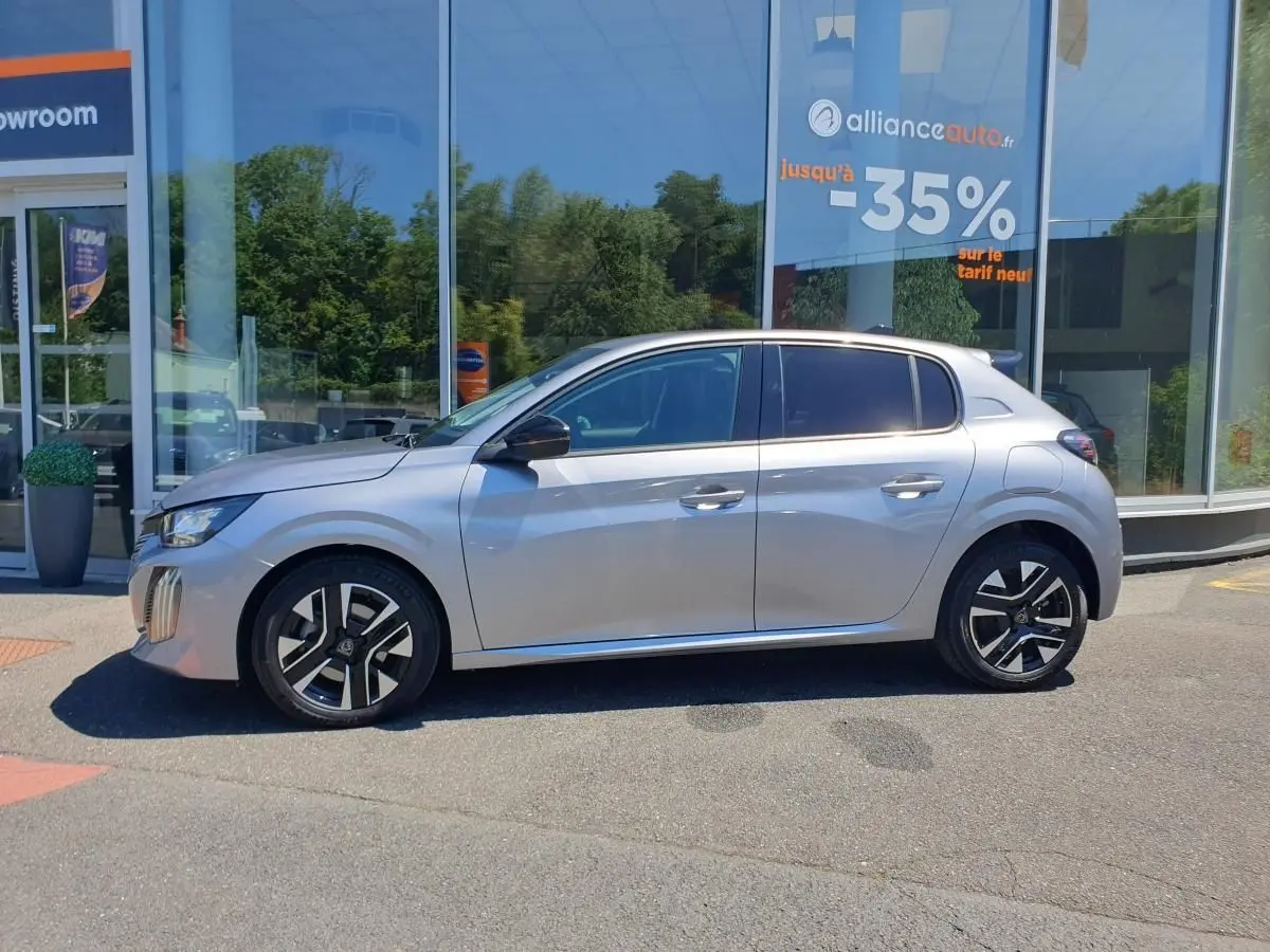 Profil droit de la Peugeot 208 1.2 Hybrid 110 Allure gris Artense avec jantes alliage 16'' bi-tons devant un showroom.