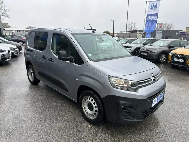 Citroën Berlingo van gris métal vu de trois quarts avant droit sur parking humide avec phares allumés.