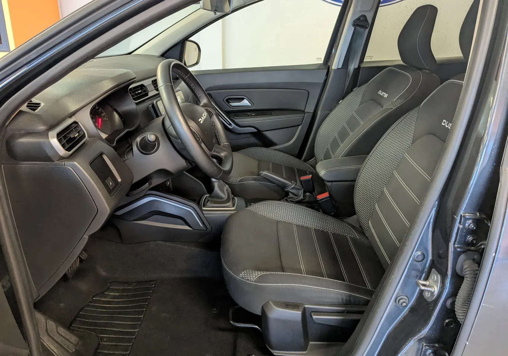Vue intérieure côté conducteur du Dacia Duster gris 2023, avec sièges noirs marqués Duster et volant multifonction.
