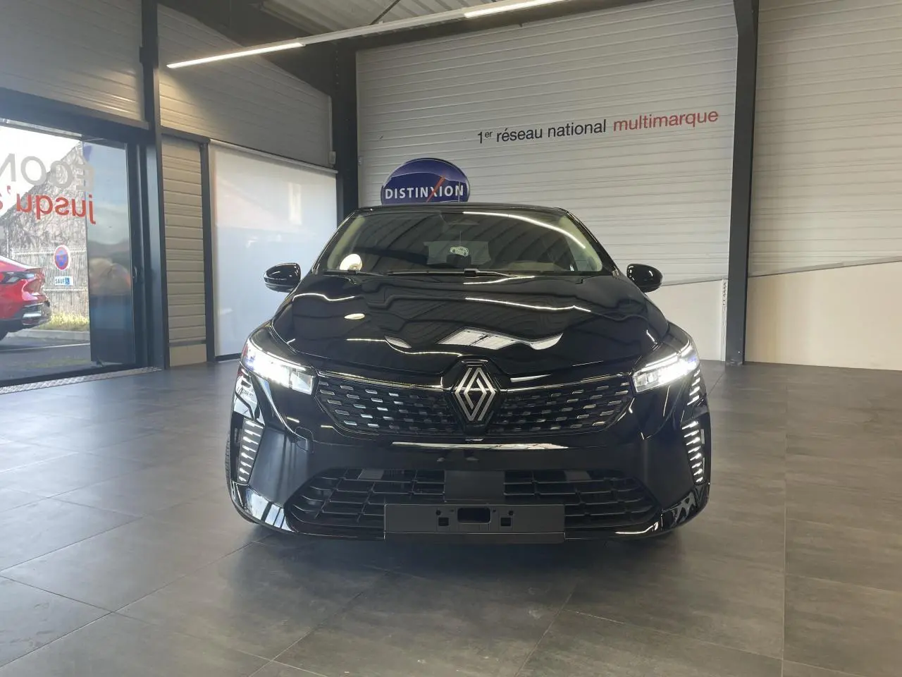 Vue de face d'une Renault Clio 1.5 Blue dCi 2025 en noir étoilé avec calandre et feux LED allumés dans un showroom.