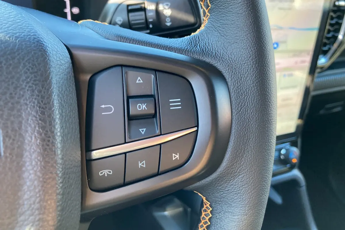 Gros plan sur les commandes multifonctions en cuir noir du volant du Ford Ranger Wildtrak blanc glacier, avec écran tactile en arrière-plan.