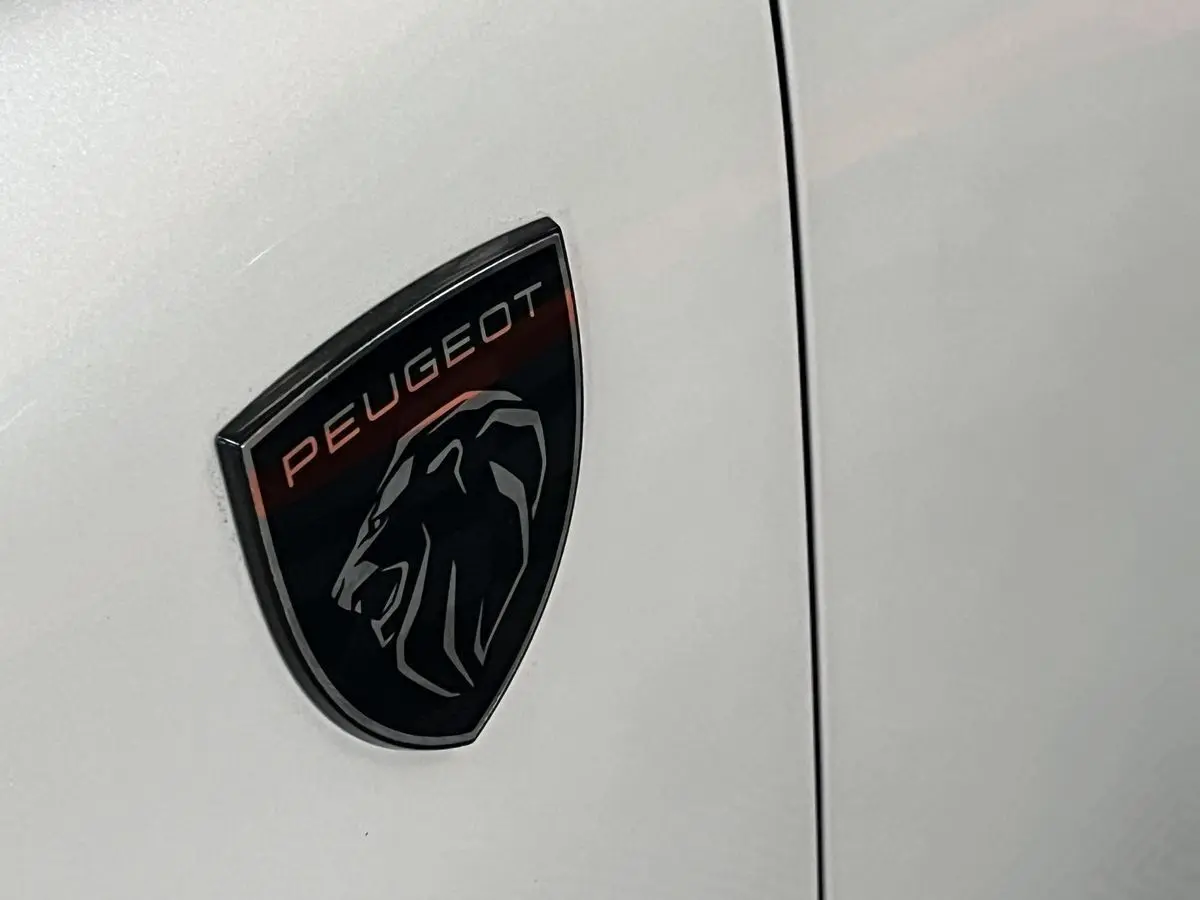 Gros plan sur l'écusson Peugeot noir et rouge sur la carrosserie blanc nacré d'une 308 GT PHEV.