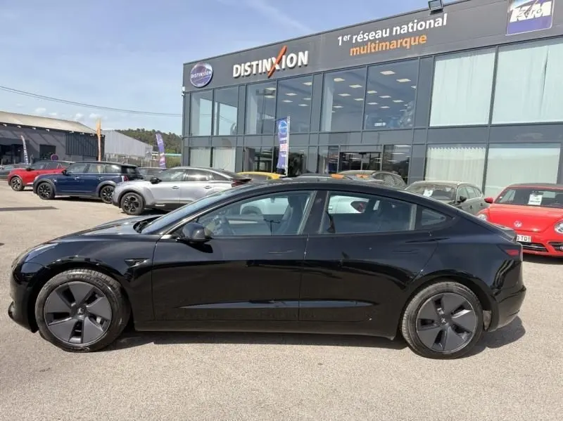 Tesla Model 3 Standard RWD Plus noire vue de profil côté gauche, avec jantes aérodynamiques, stationnée devant un showroom.