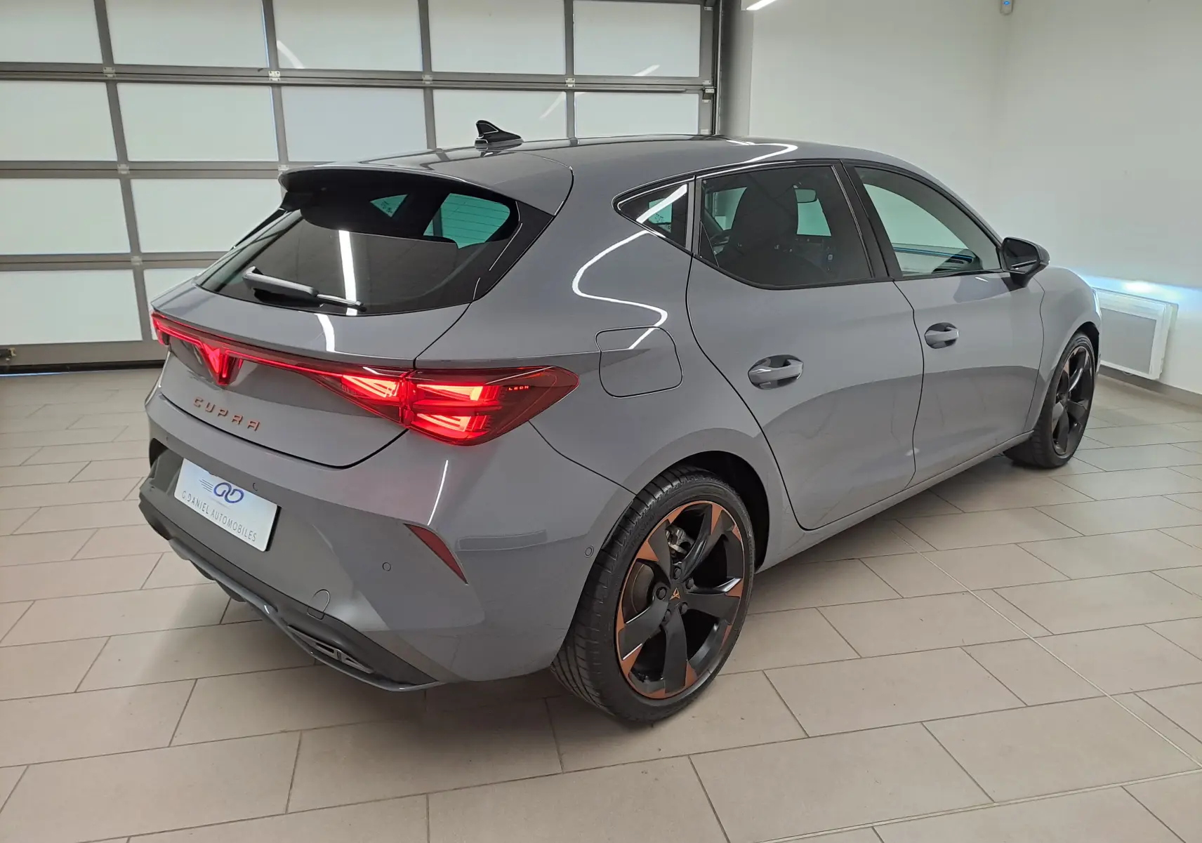 Vue 3/4 arrière droite d'une CUPRA Leon 2025 gris Graphène avec jantes cuivrées et toit ouvrant panoramique en intérieur.