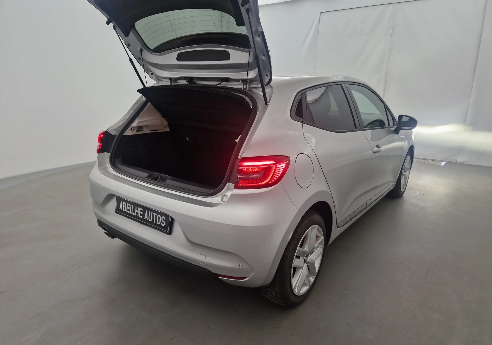 Renault Clio V gris platine vue 3/4 arrière droit avec coffre ouvert et feux arrière allumés dans un studio blanc.