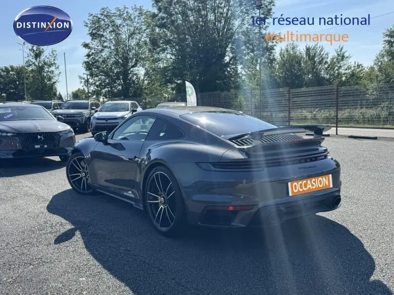 Vue 3/4 arrière droite d'une Porsche 911 Turbo S grise métallisée avec becquet arrière déployé sur parking.