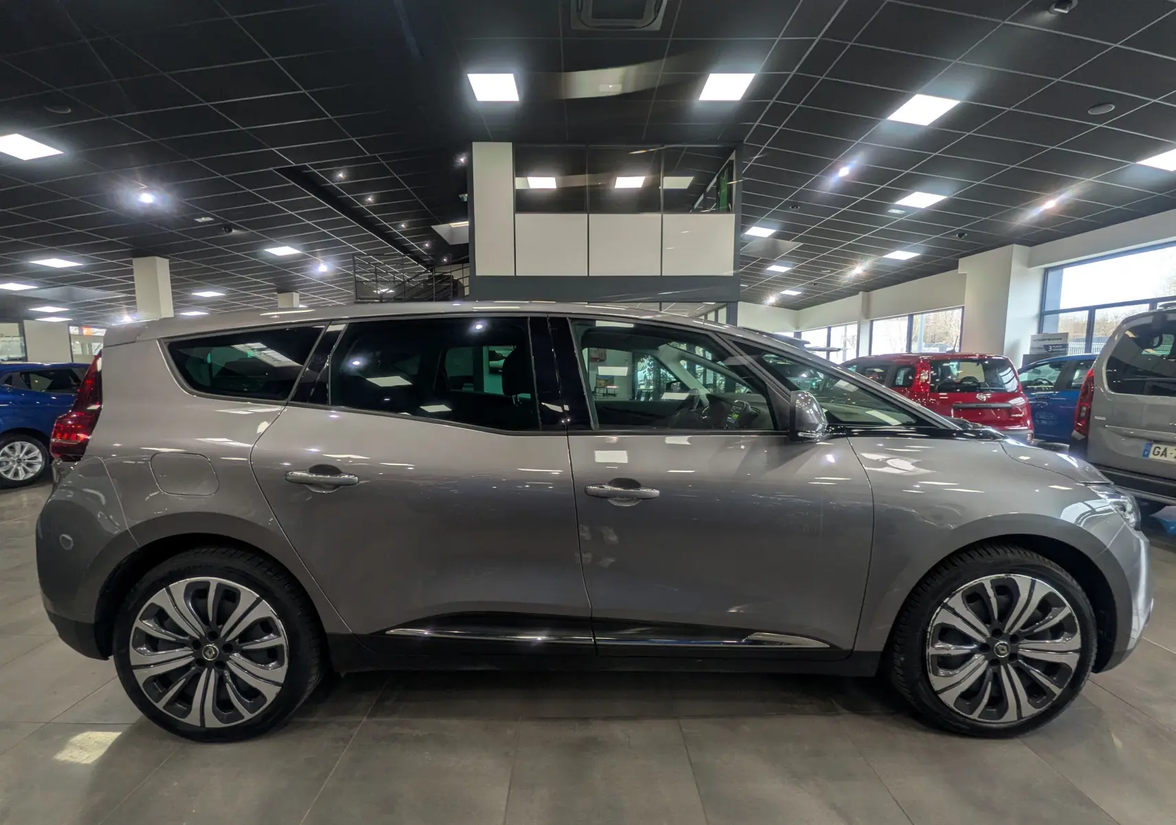 Vue de profil côté gauche d’un Renault Grand Scenic IV gris Cassiopé en showroom, avec jantes alliage distinctives.